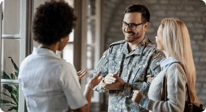 A person in a military uniform smiles while talking to a woman and a healthcare worker in a bright room.