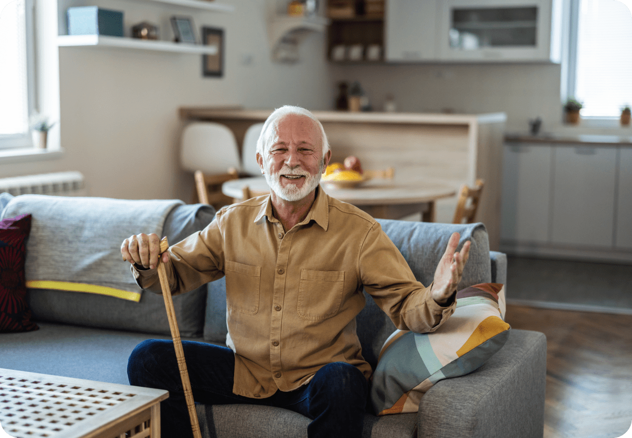 Elderly man with white hair and beard smiles while sitting on a sofa, holding a cane in a cozy living room.