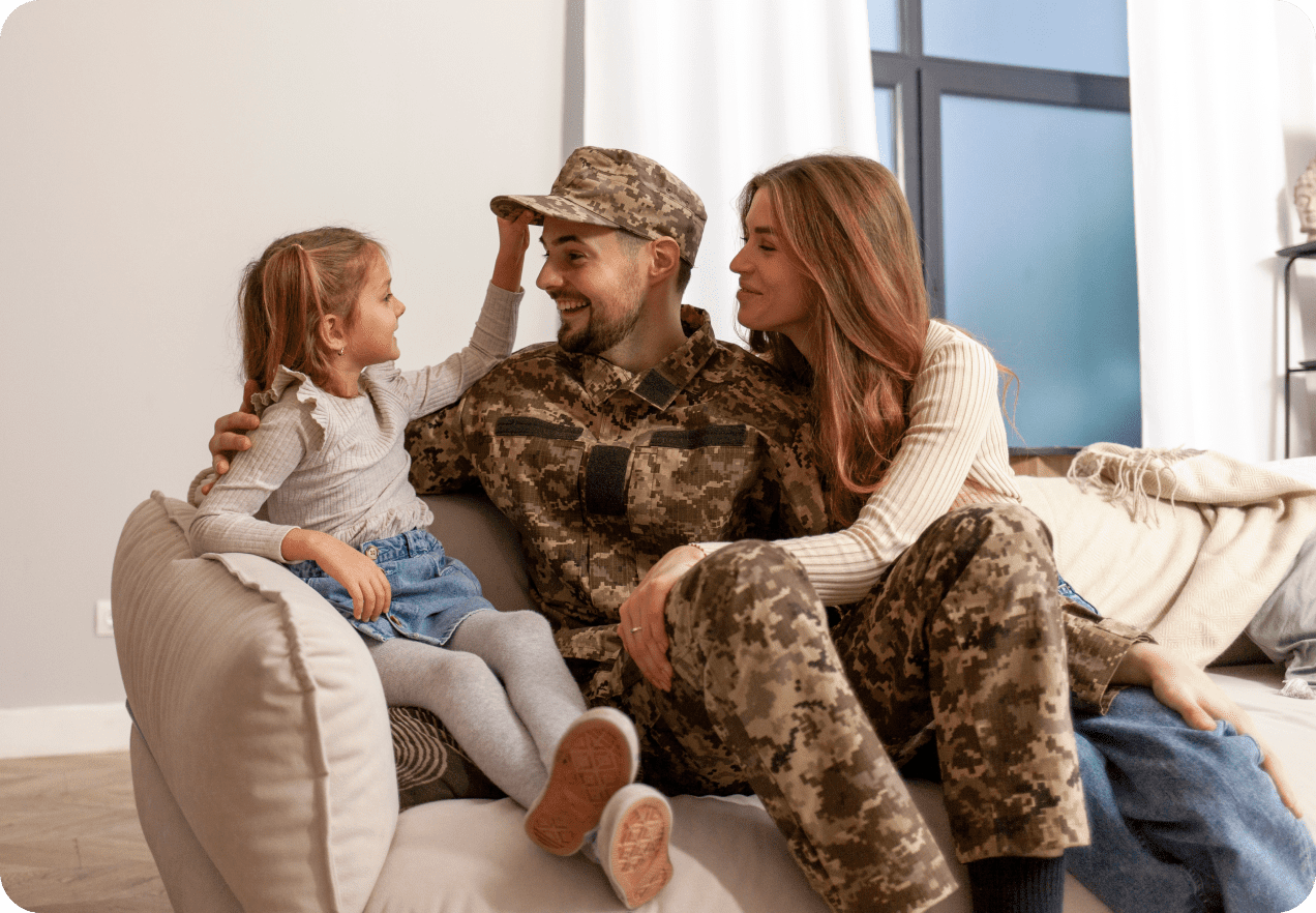 A man in military uniform sits on a couch with a woman and young girl. The girl touches the man's cap. The woman smiles at the girl. They are in a well-lit living room.