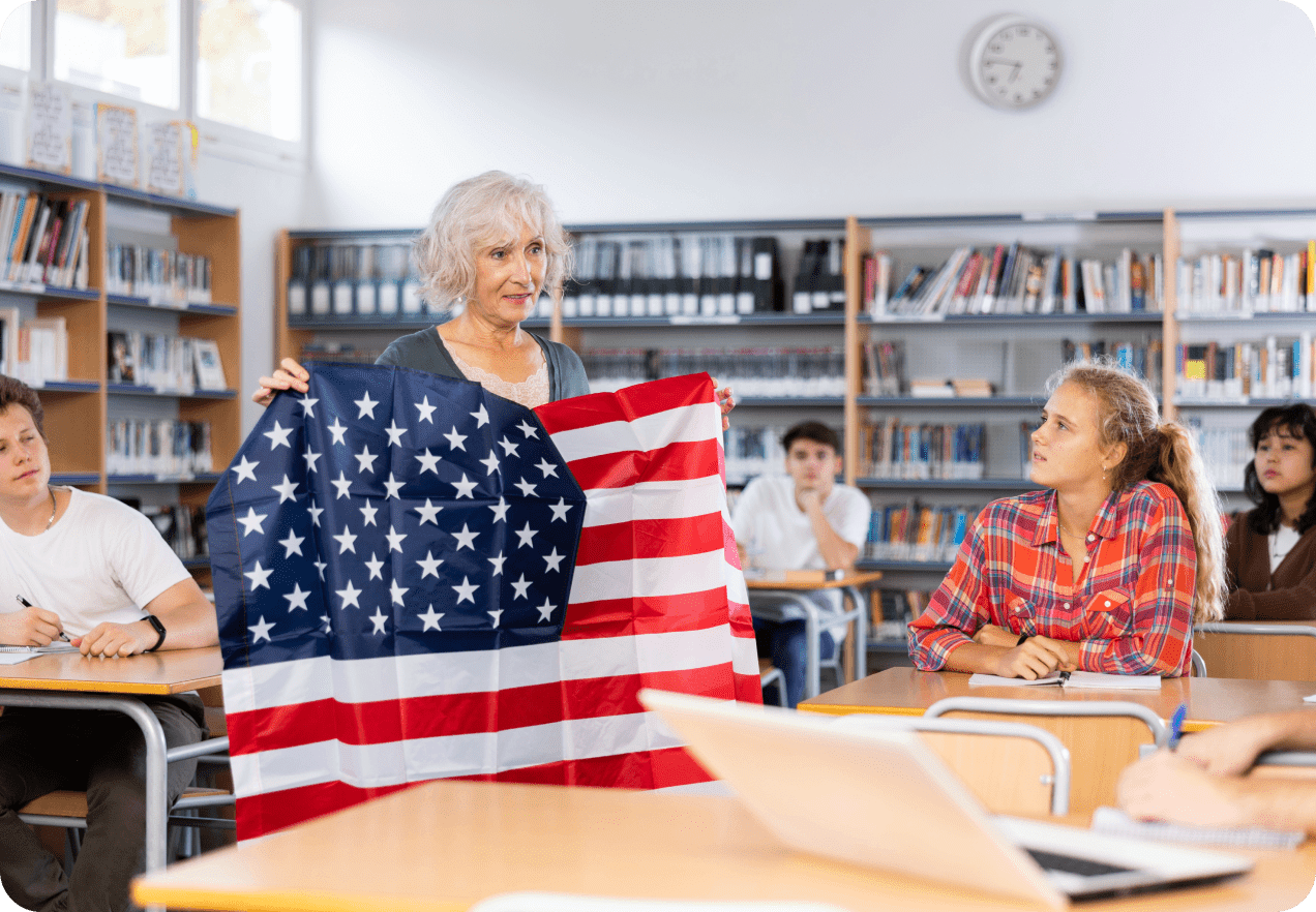 Teacher holding a U.S. flag in a classroom while students seated at desks listen. Bookshelves and a wall clock are visible in the background.
