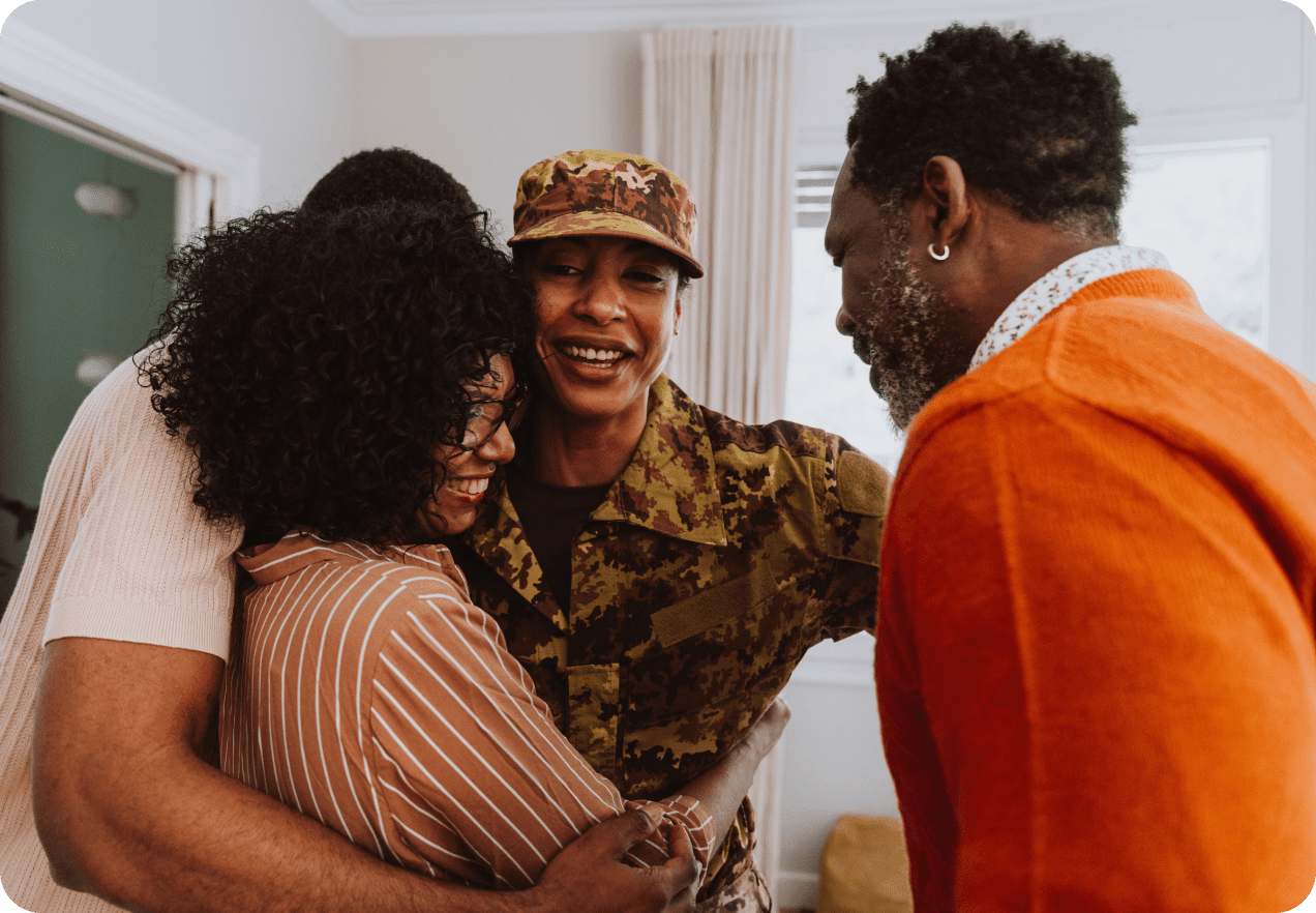 A person in a military uniform is smiling and hugging three people in a cozy, well-lit room.