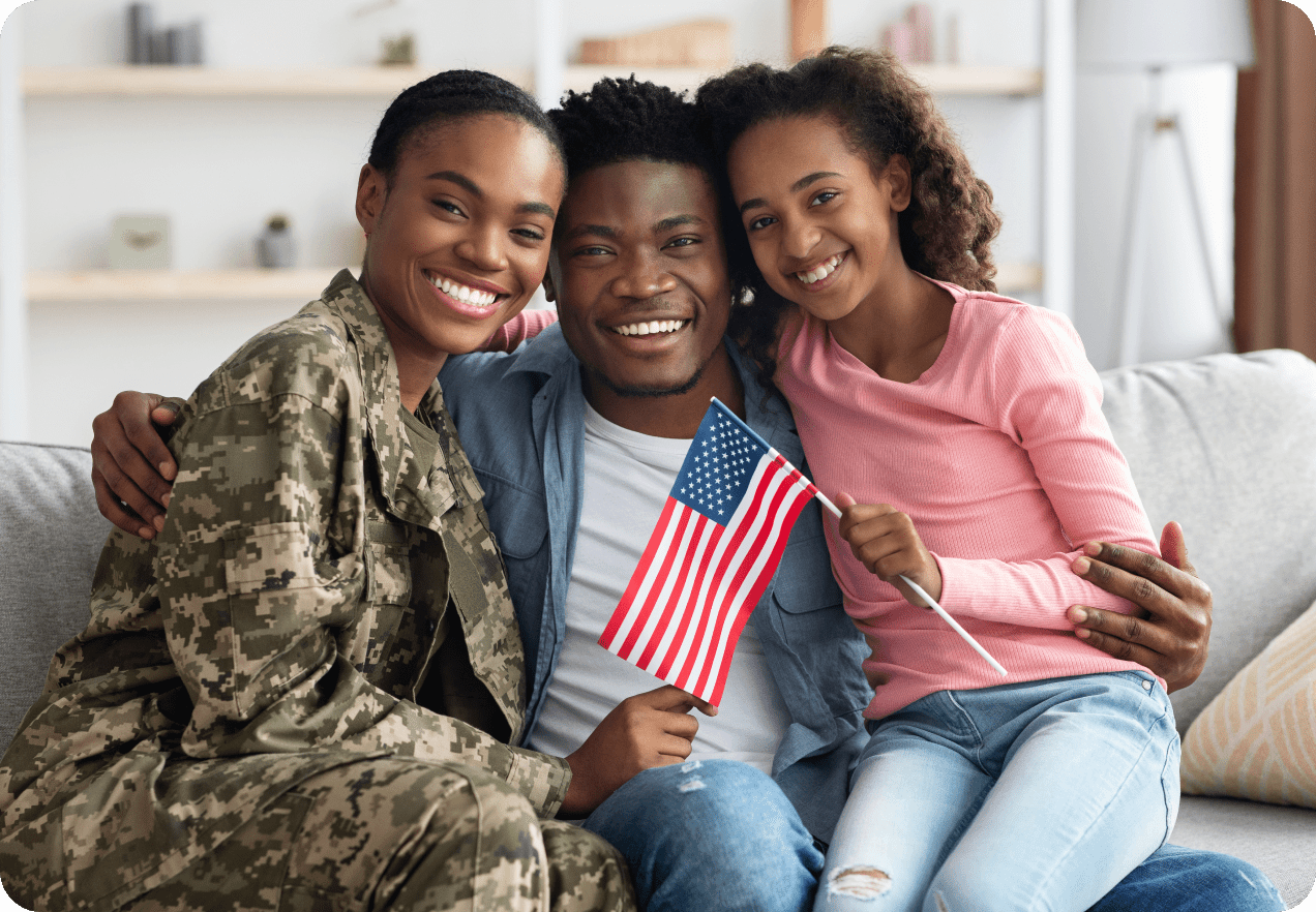 A smiling family sits on a couch, with one person in military uniform holding an American flag alongside another person holding a smaller flag.