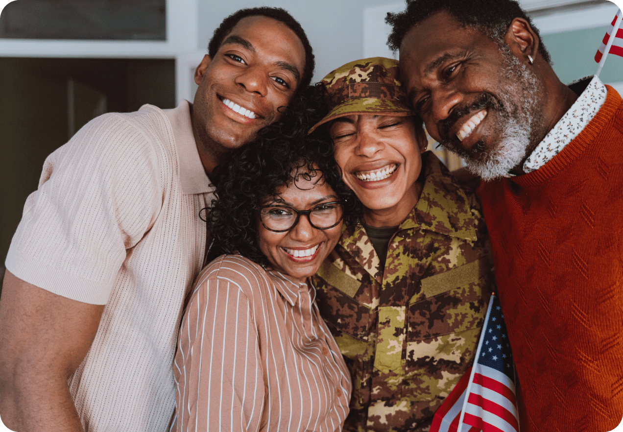 Four people smiling; one in military camouflage, holding an American flag. They stand closely together indoors, appearing happy and united.