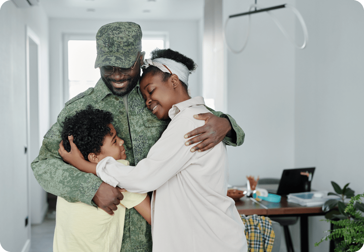 A person in a military uniform hugs two smiling children in a bright room. A table with a laptop and food is in the background.