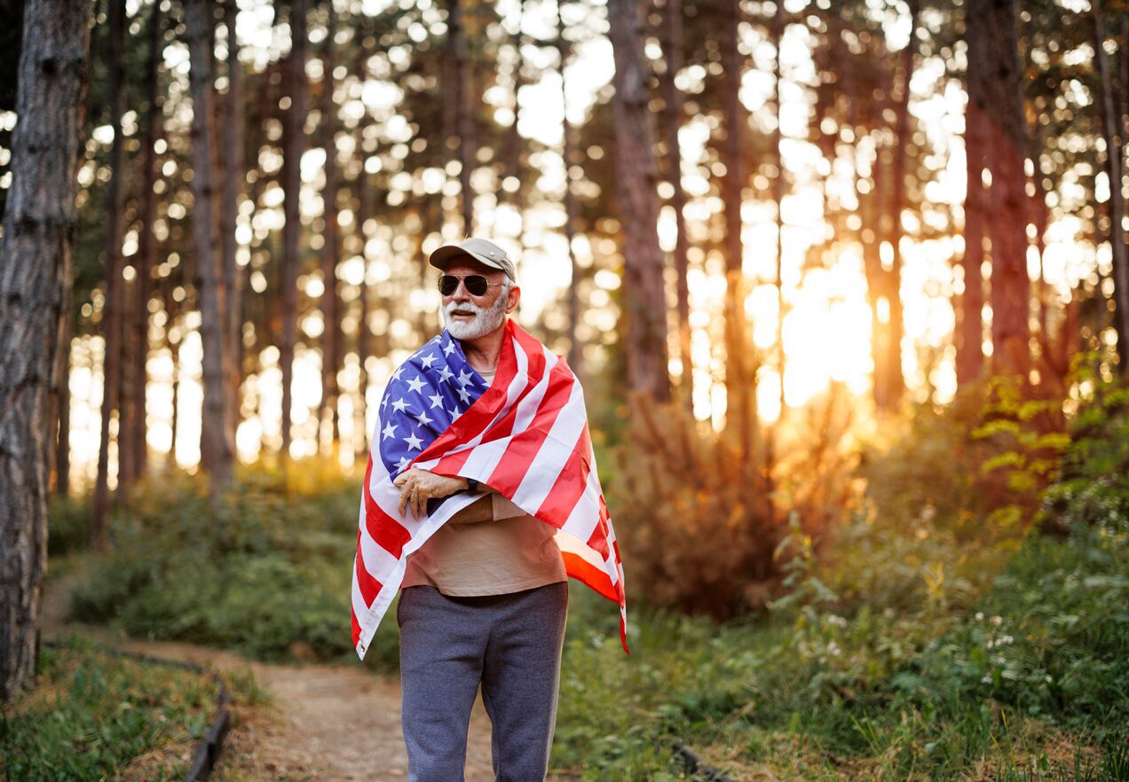 GettyImages-2179764885 A man holding an american flag in the woods.