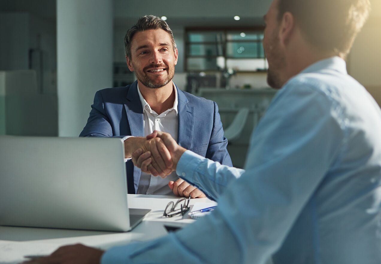 GettyImages-540525954 Two men shaking hands over a table with a laptop.
