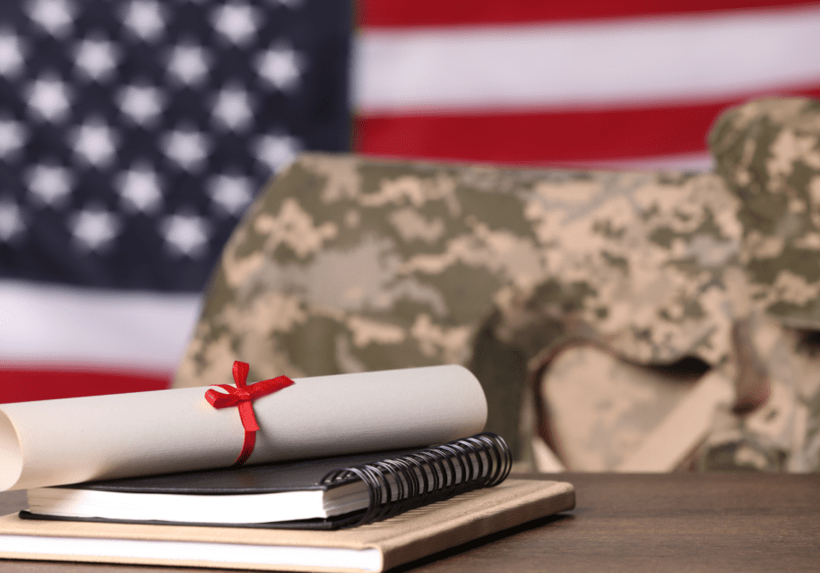 A rolled diploma with a red ribbon on notebooks sits in front of a camouflage uniform and a blurred American flag.